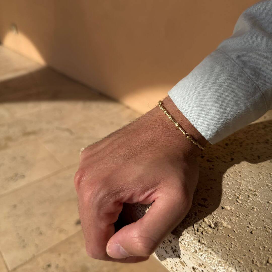 Close-up of the Breindell Tzitzit I Bracelet in 14K gold, worn on a man’s wrist with a light blue fitted shirt cuff. The hand is resting on a textured stone bench, highlighting the architectural geometric links and polished gold finish. High-end Israeli jewelry combining modern design with sacred Jewish symbolism.