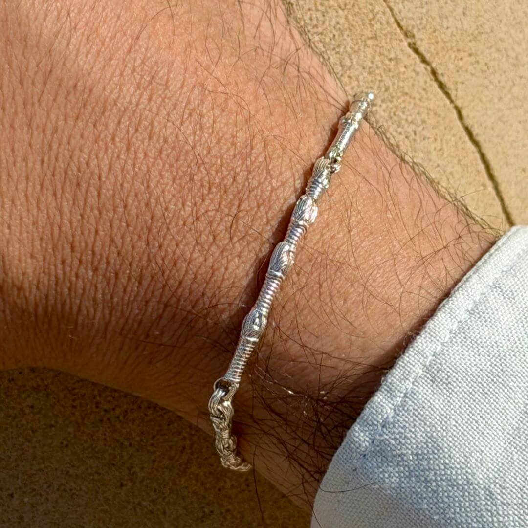 Tzitzit I Bracelet in Sterling Silver; premium Jewish jewelry made in Israel. Close-up on a man’s hand with a light blue cuff against a stone background. This architectural unisex bracelet features symbolic winding links in a polished 925 silver finish.
