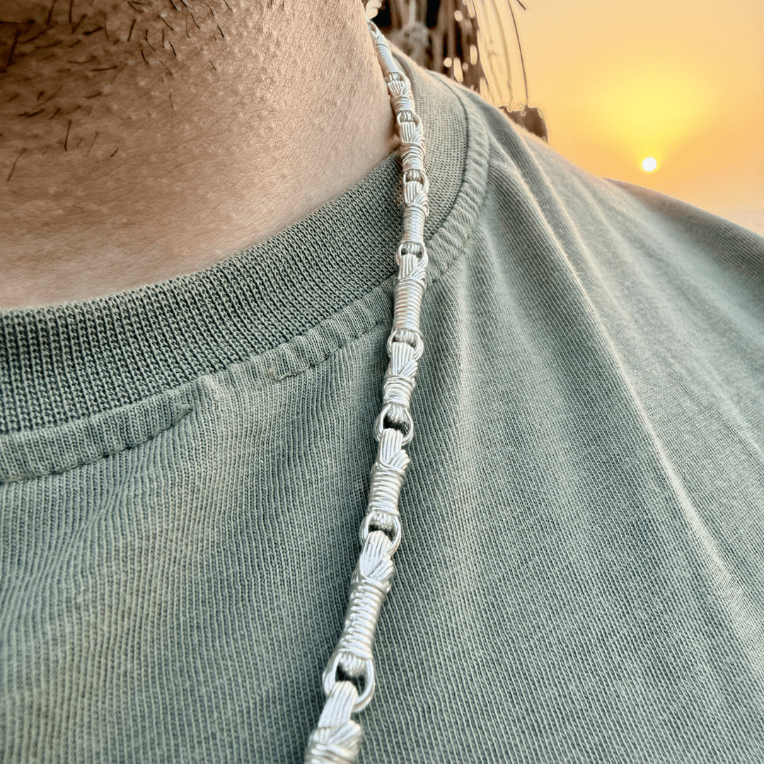 Close-up of the Breindell Tzitzit I Necklace №2 in 925 Sterling Silver, worn by a male model over a khaki green t-shirt. The polished architectural pendant, featuring the 7, 8, 11, and 13 winding links, is showcased in a lifestyle setting with a blurred Israeli beach and palm trees in the background. High-end, minimalist Jewish jewelry handcrafted in Israel.