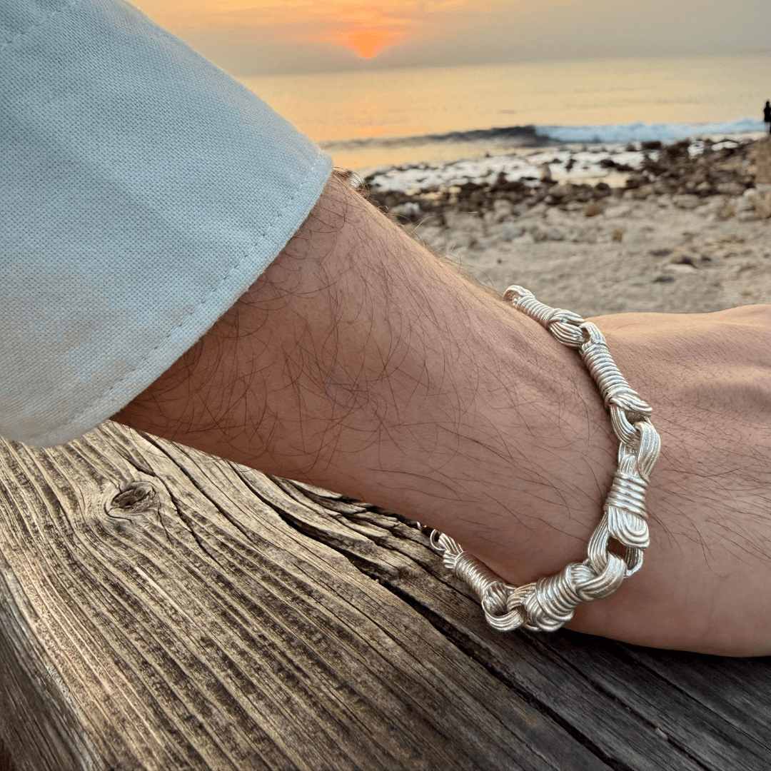 Lifestyle view of the Breindell Tzitzit I Bracelet №3 in 925 Sterling Silver during sunset. Worn on a wrist leaning against a weathered wooden boardwalk ramp, the Mediterranean Sea and Israeli coastline are blurred in the background. The jewelry features seven bold and chunky, handcrafted architectural segments in a polished modernist finish.