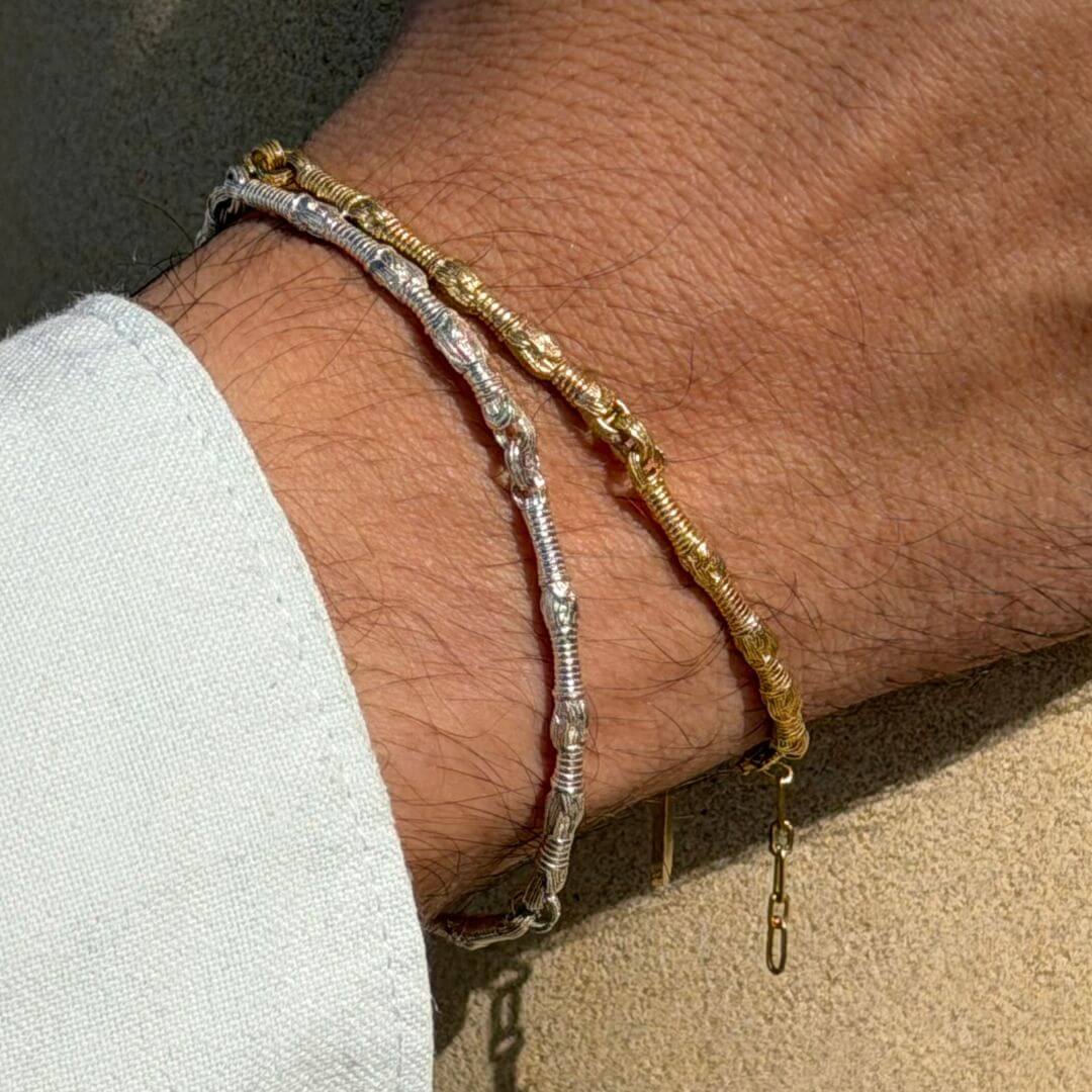 Lifestyle close-up of a man’s wrist wearing the Breindell Tzitzit I Bracelet in 925 Sterling Silver and the matching version in 14K Gold. The mixed-metal stack highlights the shared architectural link design and symbolic windings against a baby blue shirt cuff and textured stone pillar.