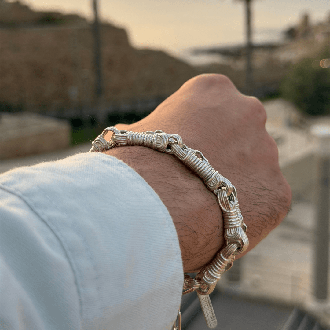 Close-up of stacked Breindell jewelry: the bold Tzitzit I Bracelet №3 in 925 Sterling Silver with the intricate №2 nestled behind it, with blurred palm trees and a Mediterranean sunset in the background. 
