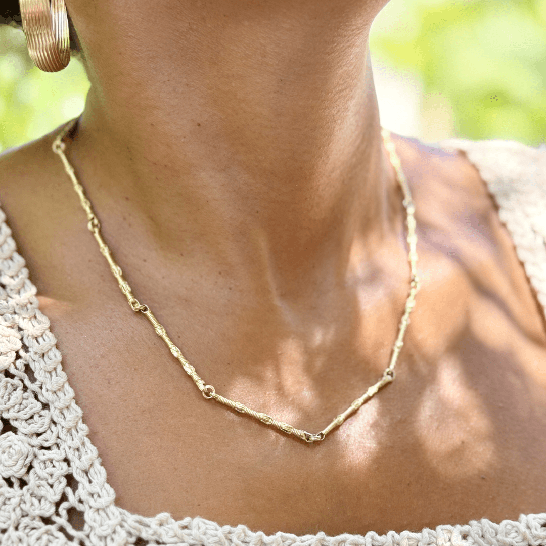 Female model wearing the 14K Gold Breindell Tzitzit I Necklace №1 and gold hoop earrings, styled with a cream crochet top against a background of green leaves