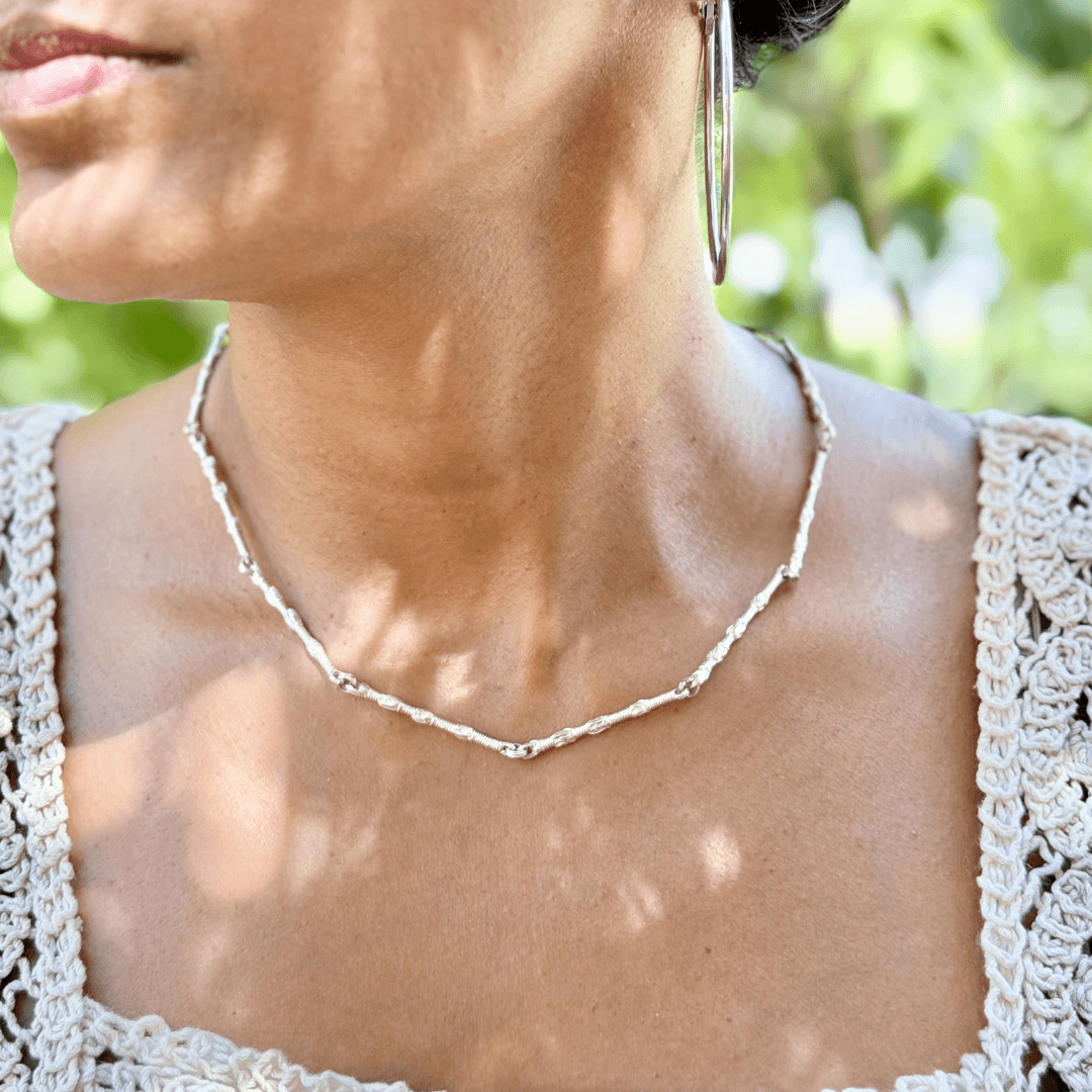 Female model wearing the Sterling Silver Breindell Tzitzit I Necklace №1, styled with a cream crochet top against a background of trees with green leaves.